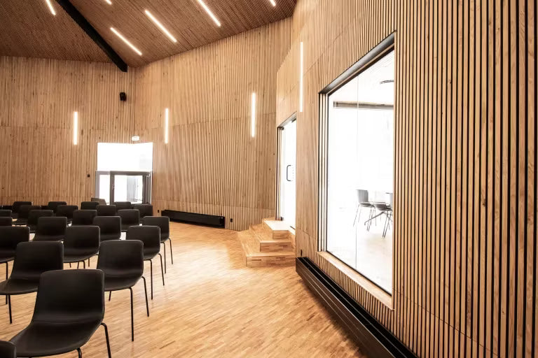 Spacious conference room with Akupanel acoustic wood panels on walls and ceiling, rows of black chairs, large windows, and vertical light fixtures.